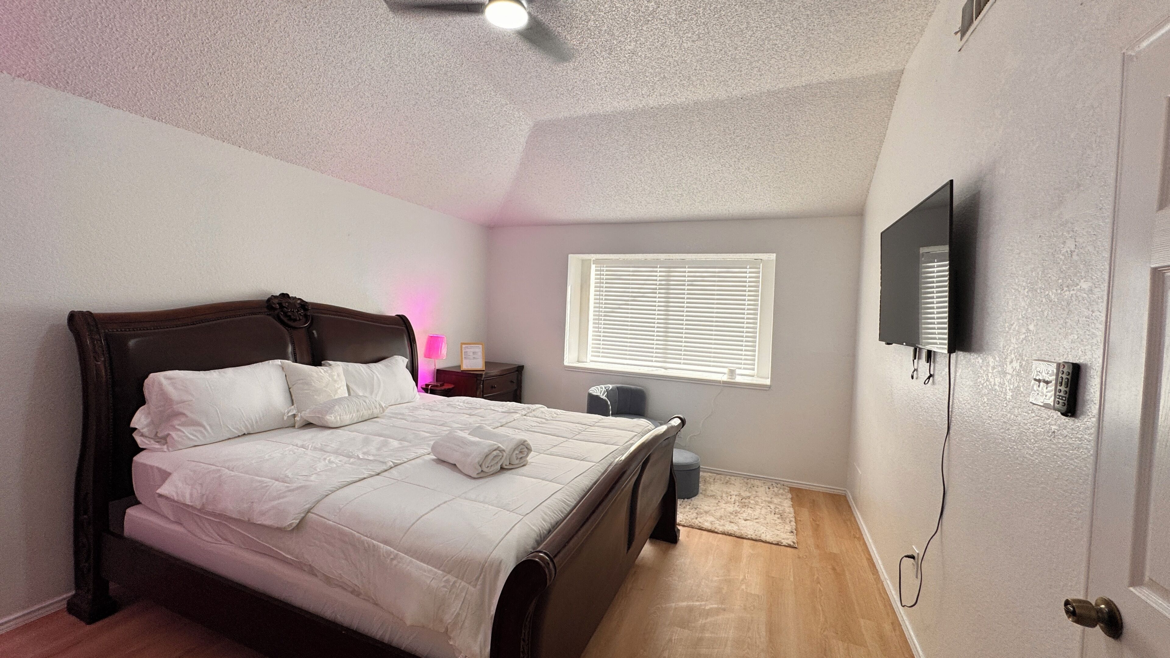 A bedroom with a bed, bedside table, lamp, window, and a TV mounted on the wall.