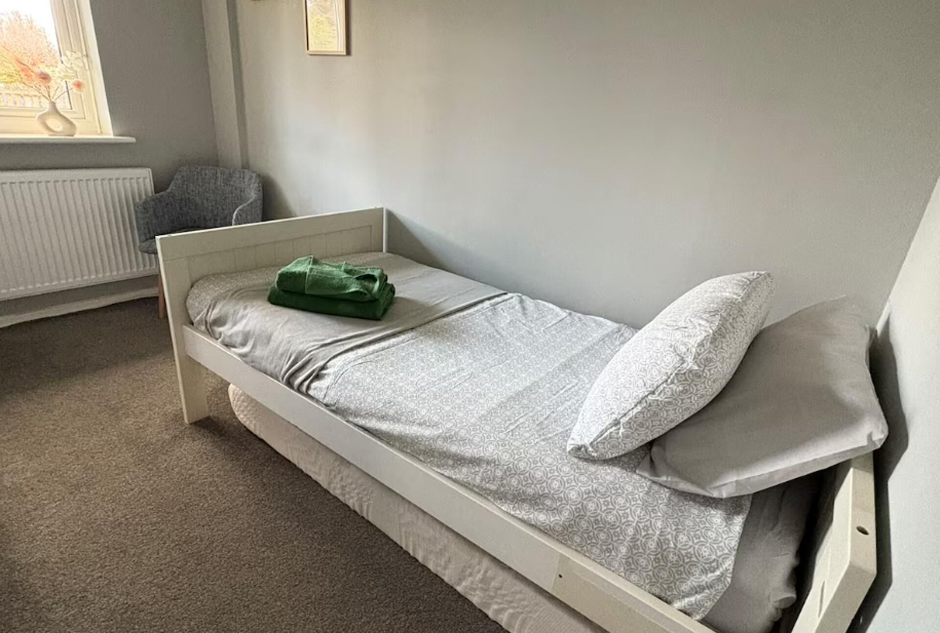 A single-bed room with a white frame, grey bedding, and a window with a view of trees.