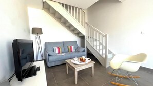 Ein modernes Wohnzimmer mit einem grauen Sofa, einem Couchtisch mit Kuchen, einem Fernseher, einer Lampe und einem Schaukelstuhl.