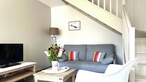 Un salon moderne avec un canapé gris, un fauteuil blanc, une table basse, un téléviseur et un escalier.