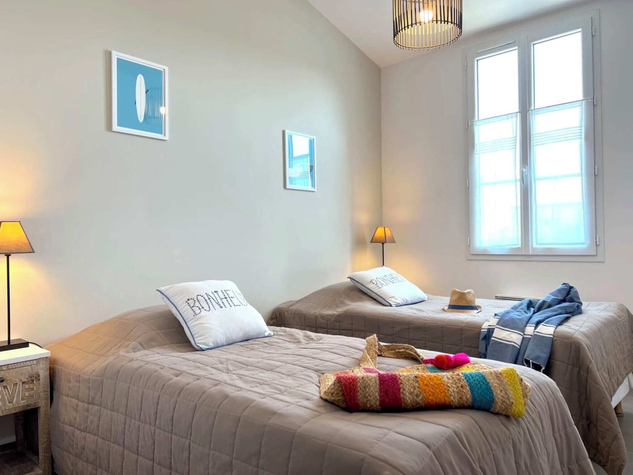 Une chambre d’hôtel avec deux lits, une fenêtre avec des stores, une lampe et un mur orné de deux tableaux encadrés.