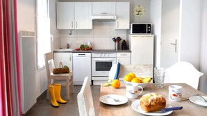 A compact kitchen with white cabinets, a microwave, and a stove. There is a dining table with a loaf of bread, a bowl of fruit, and a cup. - Villa n°8 - The little sergeant (La Flotte)
