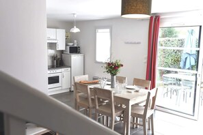 A modern kitchen with white appliances, a dining area with wooden chairs and a table, and a view of the outdoors through a sliding glass door. - Villa n°13 - Le lizay (La Flotte)