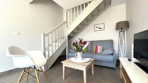 Una sala de estar moderna con una escalera, un sofá gris, una silla blanca, una mesa de centro con un jarrón floral, un televisor y una lámpara de pie.