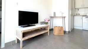 A modern living room with a flat-screen TV on a wooden console table, a small side table with a vase of flowers, and a kitchen area with white cabinets and a dishwasher. - Villa n°6 - The Target (La Flotte)