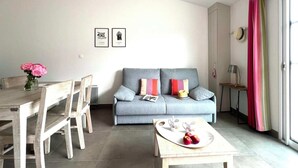 A modern living room with a grey sofa, wooden dining table, and chairs. There are two framed pictures on the wall, a vase with flowers on the table, and a tray with apples and cups on a small table. - Villa n°6 - The Target (La Flotte)