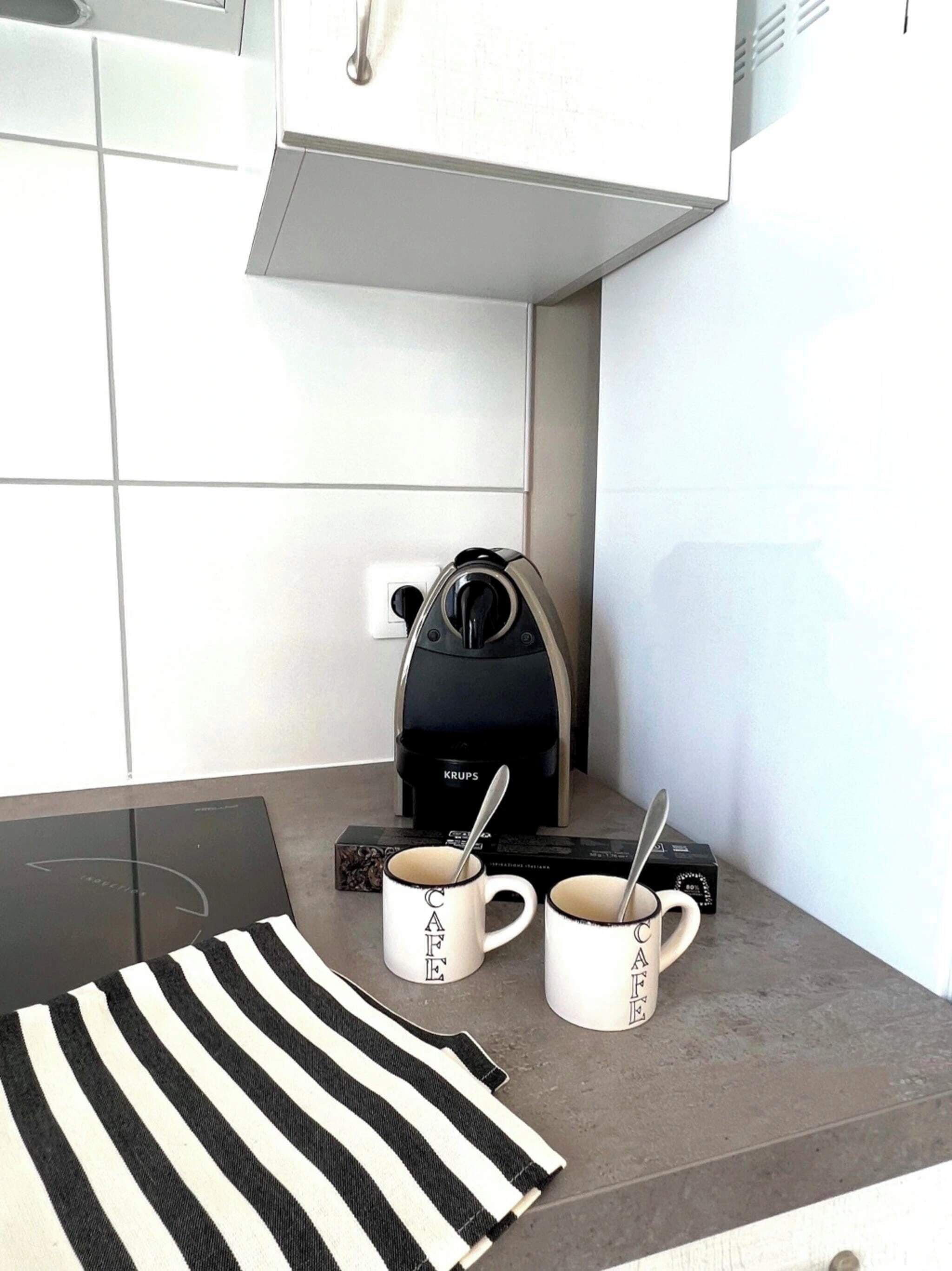 Une cuisine équipée d’une machine à café, de deux tasses et d’un torchon à rayures posé sur le comptoir.
