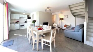 A modern kitchen with white cabinets, a dining table with chairs, a grey sofa, and a staircase. - Villa n°5 - Les gollandières (La Flotte)