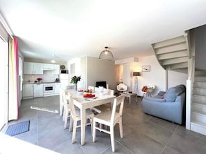 A modern kitchen with white cabinets, a dining table with chairs, a grey sofa, and a staircase. - Villa n°5 - Les gollandières (La Flotte)