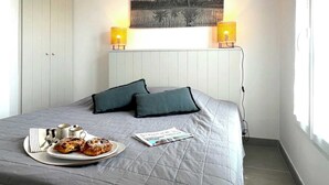 A neatly made bed with a grey quilt, two pillows, a plate of pastries, and a magazine on a bedside table. There are two wall-mounted lamps and a painting above the bed. - Villa n°5 - Les gollandières (La Flotte)