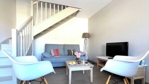 A modern living room with a staircase, a grey sofa, two white chairs, a coffee table, a TV, and a floor lamp. - Villa n°9 - Le martray (La Flotte)