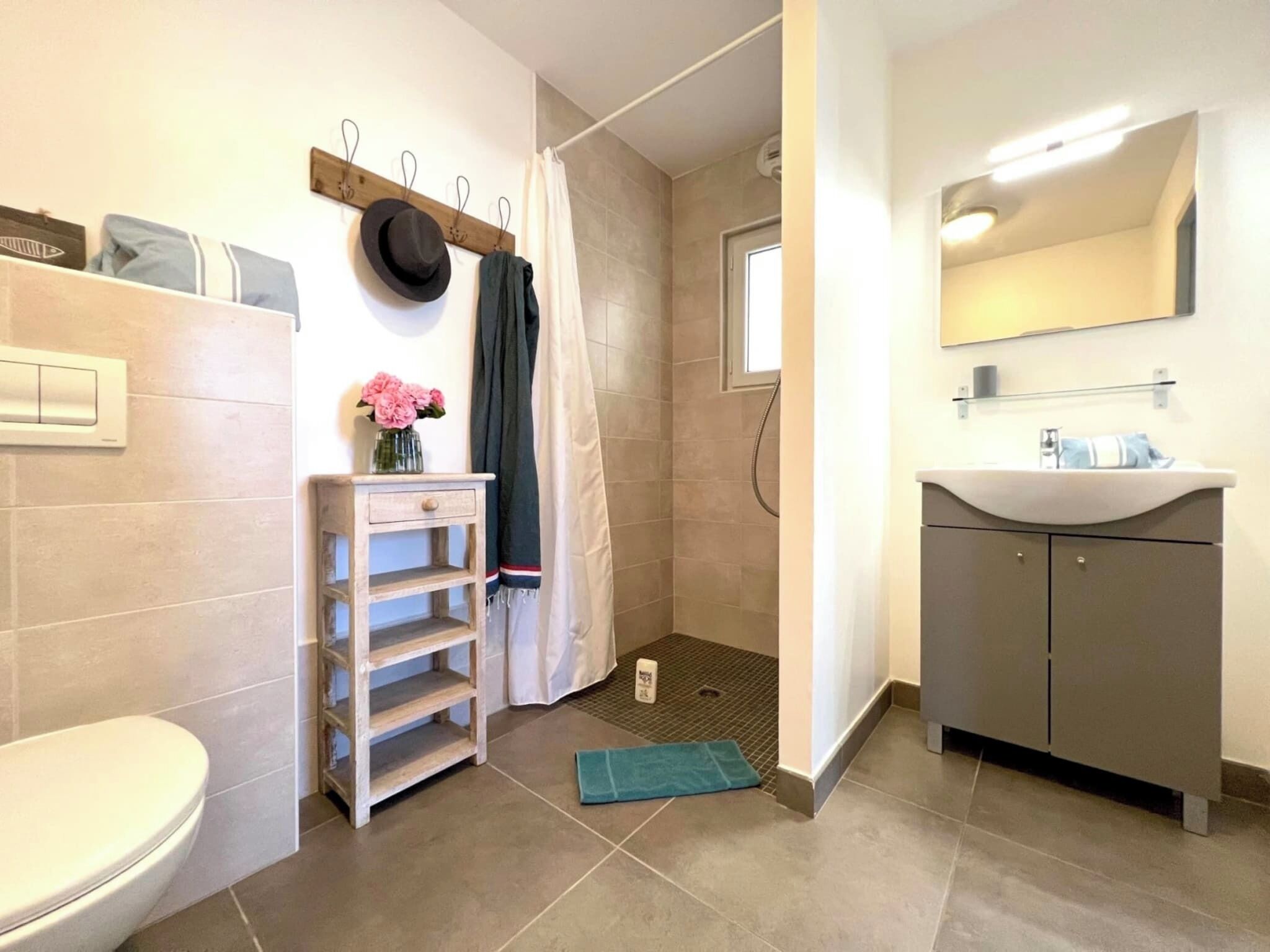 A bathroom with a toilet, a sink, a shower, and a towel rack with hats and a vase of flowers.