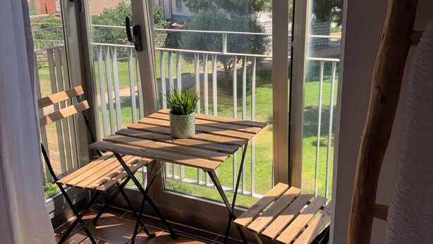 Un balcón con una mesa y sillas de madera, una planta en maceta y vistas a un césped verde y edificios a lo lejos.