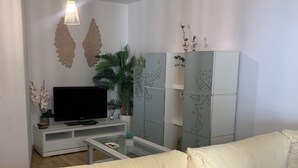 Una sala de estar moderna con un sofá, un televisor de pantalla plana, una mesa de centro de vidrio, plantas decorativas y un mueble blanco con motivos florales.