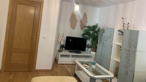 Una sala de estar con puerta de madera, un televisor de pantalla plana sobre un soporte blanco, una mesa de centro de vidrio, decoración mural y una lámpara de techo.