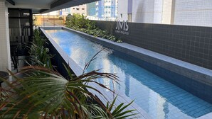 A swimming pool with a tiled edge, surrounded by a low wall with greenery. A sign reading "JMS CONSTRUÇÕES" is visible on the wall. - Flat Quality Comfort CoHouse - Tambaú (João Pessoa)