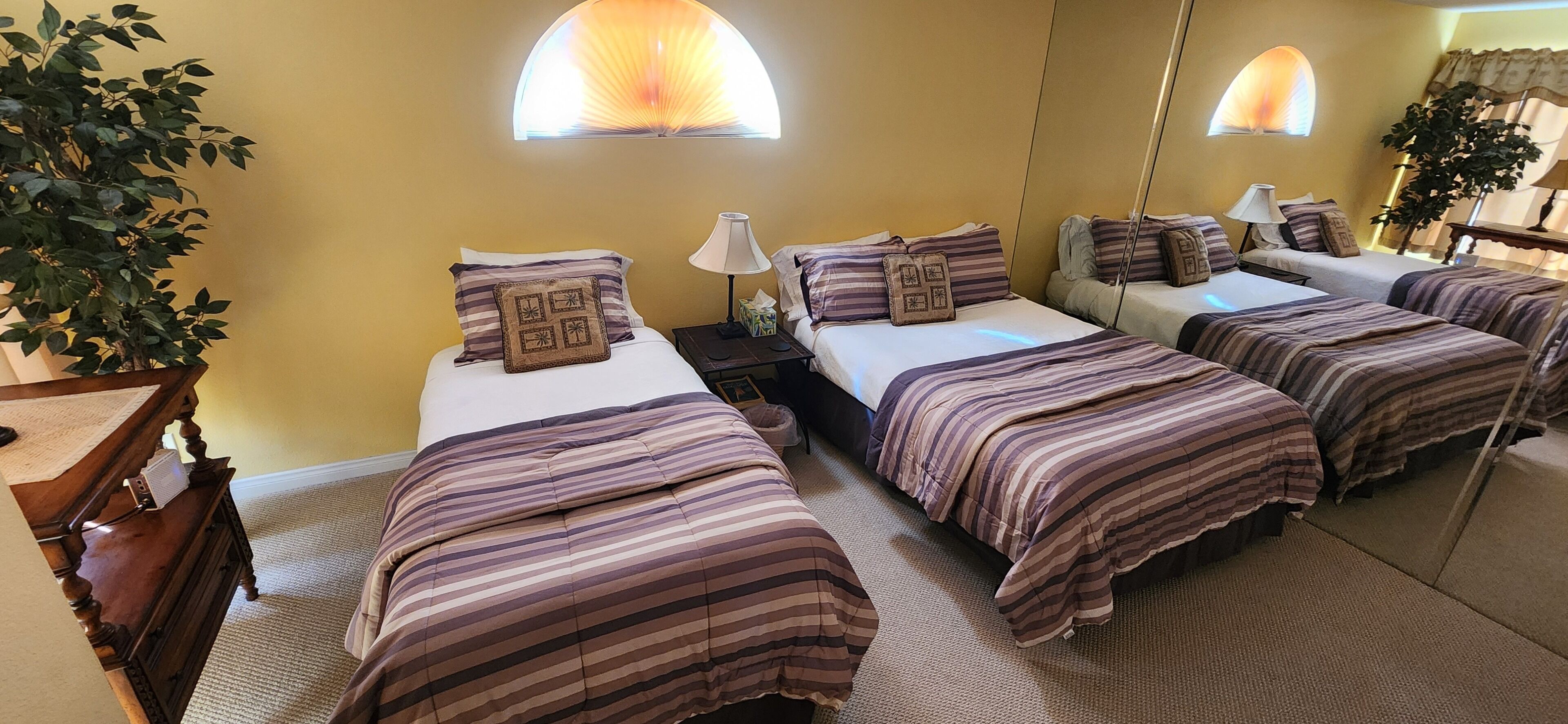 Quarto de hotel com quatro camas, cada uma com roupa de cama listrada e almofadas combinando. Há duas mesas de cabeceira com abajures, um espelho grande e um vaso de plantas.