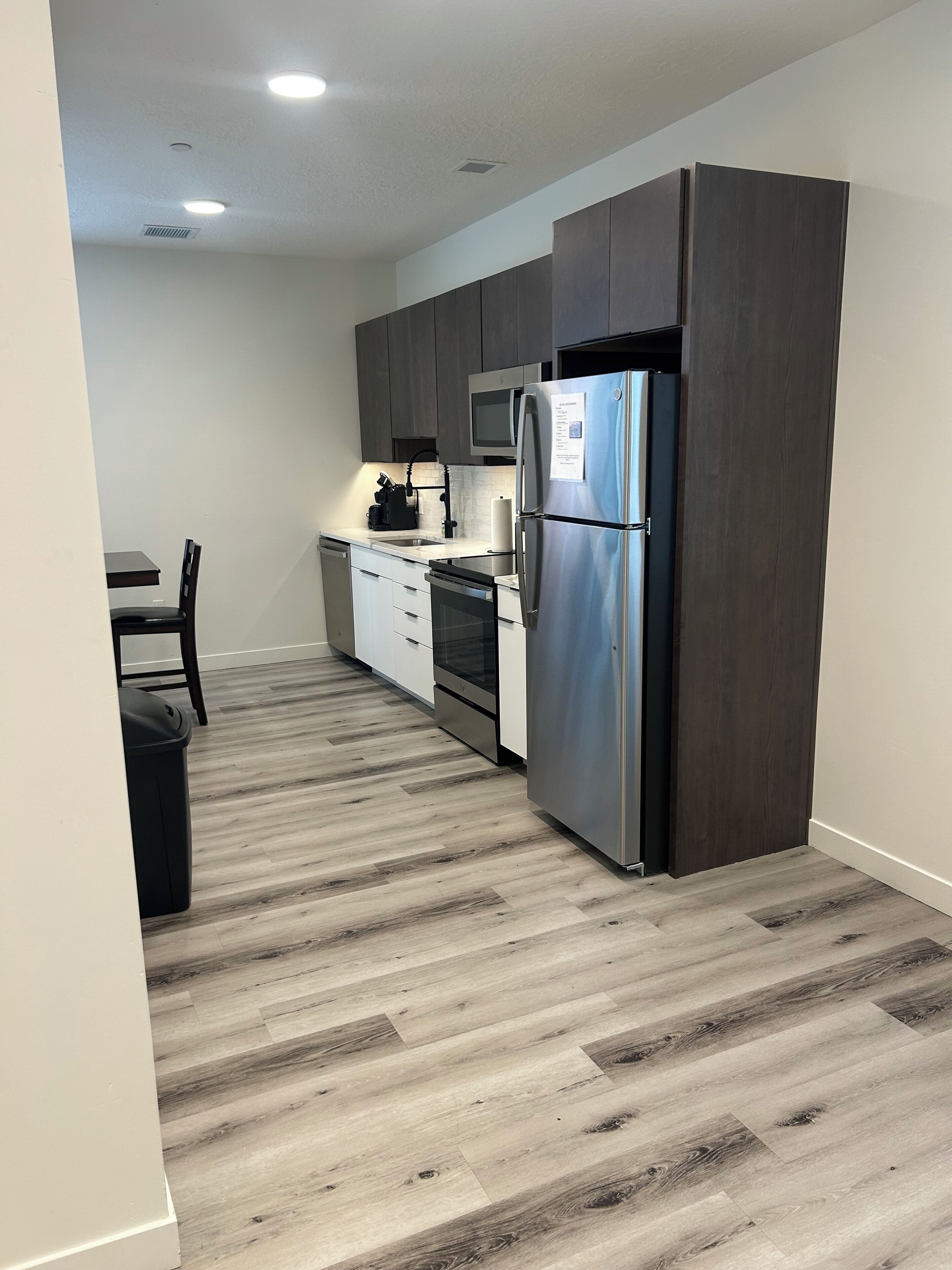 A modern kitchen with dark cabinets, stainless steel appliances, and a wooden floor.