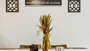 Una mesa con un jarrón de plantas secas, una escultura negra y una bandeja blanca. Dos paneles decorativos con un motivo geométrico y un cartel que dice: "Buena comida. Buenos amigos. Buenos momentos."
