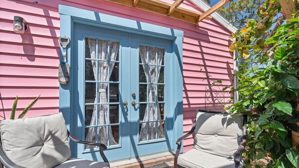 Une maison rose avec une porte bleue, deux chaises à l’extérieur et un porche en bois.