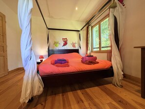Ein Schlafzimmer mit Holzboden, einem Bett mit orangefarbener Bettwäsche, einem Nachttisch mit Lampe und einem Fenster mit Vorhängen.