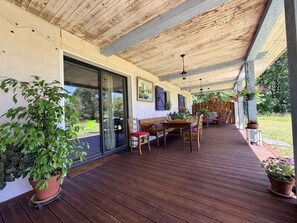 Eine überdachte Terrasse mit Holzdeck, einem Esstisch mit Stühlen, einer Couch und Topfpflanzen.