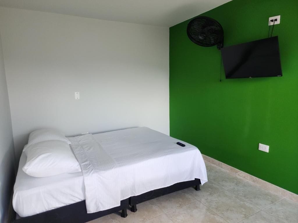 Uma cama de solteiro com roupa de cama branca num quarto com paredes verdes, um ventilador preto e uma TV montada.