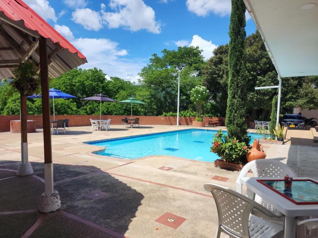 Piscina con sillones de descanso y sombrillas, rodeada de follaje y un porche cubierto.