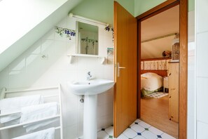 Uma casa de banho com lavatório branco, um espelho, porta-toalhas e uma porta de madeira que dá para um quarto.