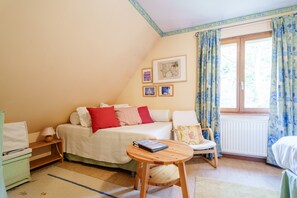 A bedroom with a bed, a chair, a small table, a nightstand, a lamp, and a window with curtains. - Darjeeling Guest Room (Boersch)