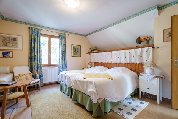 Quarto com cama, cadeira, mesa, mesa de cabeceira e janela com cortinas.