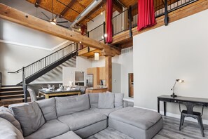 Ein modernes Wohnzimmer mit einer Eckcouch, einem Essbereich mit Tisch und Stühlen, einer Küche im Hintergrund und einer Treppe mit roten Vorhängen.