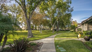 Un chemin bordé d’arbres et traversé par une pelouse bien entretenue, qui mène à un bâtiment doté d’un toit plat et d’un revêtement extérieur de couleur beige.