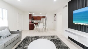 A modern living room with a grey sofa, a dining table with chairs, a black and white rug, and a large flat-screen TV mounted on a black panel. - Modern Boca Condo Near FAU : 2 KING BR + Pool (Boca Raton)