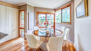 Un espace repas avec une table en verre entourée de chaises blanches, un plancher en bois et une vue sur un espace extérieur abrité.