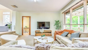 Un salon avec un téléviseur, un canapé et une table basse. Une porte donne accès à une autre pièce, et une fenêtre offre une vue sur la verdure.