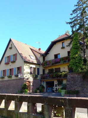 Un bâtiment de style européen traditionnel, doté d’une façade en pierre, de nombreux balcons ornés de jardinières fleuries et surplombant un ciel d’un bleu éclatant.