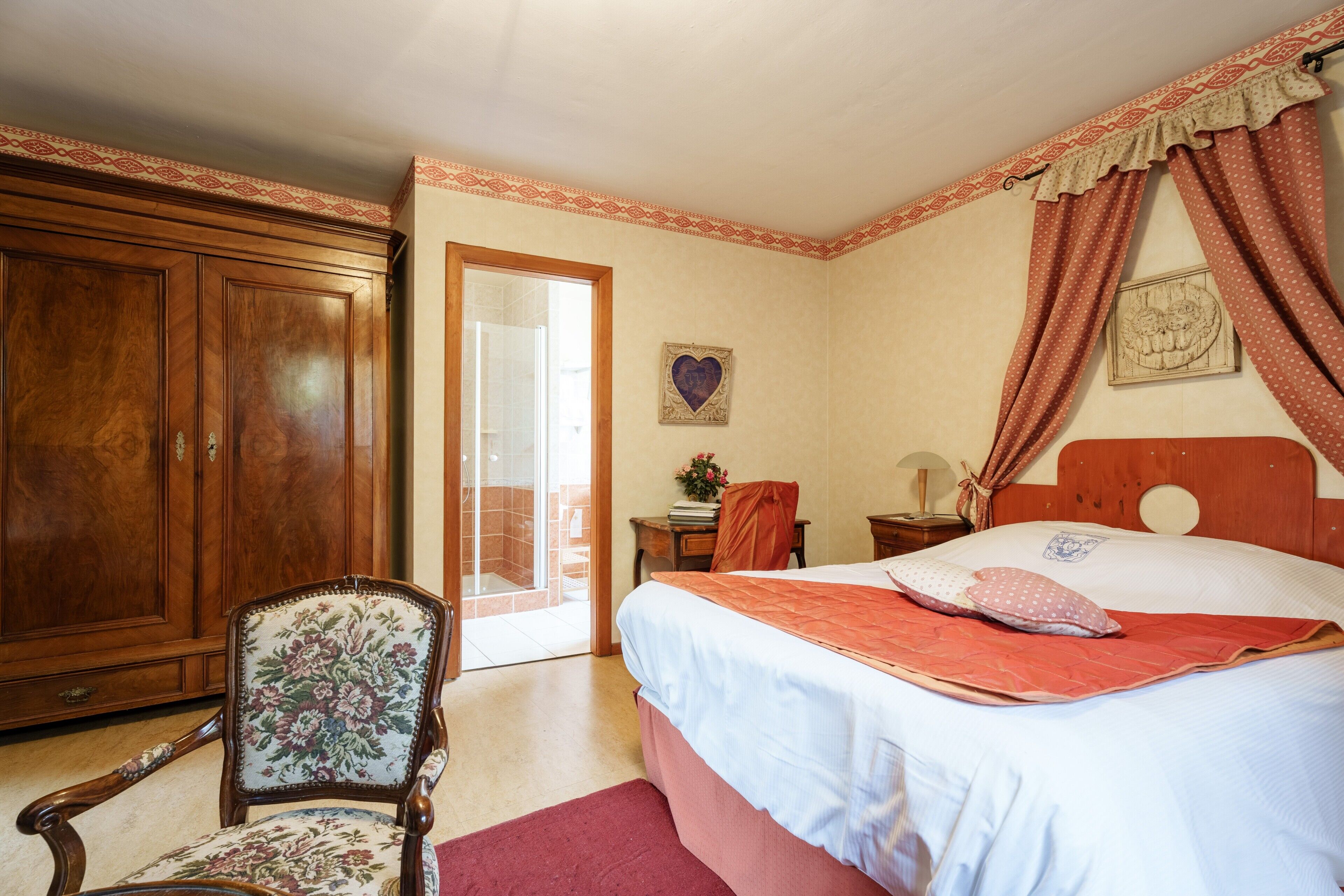 A bedroom with a large wooden wardrobe, a floral armchair, a bed with a red blanket, and a small table with a lamp.
