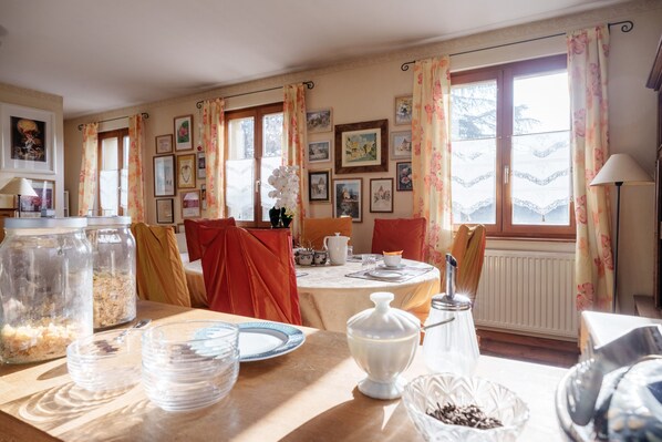 A dining area with a table set for breakfast, a window with curtains, and various framed pictures on the wall.