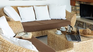 Un canapé en osier avec des coussins blancs, un ordinateur portable, une tasse et un livre posés sur un pouf tressé.