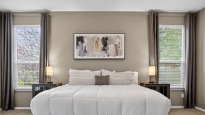 Un letto grande con piumone bianco, due comodini con lampade, un quadro astratto incorniciato sopra il letto e finestre con tende.