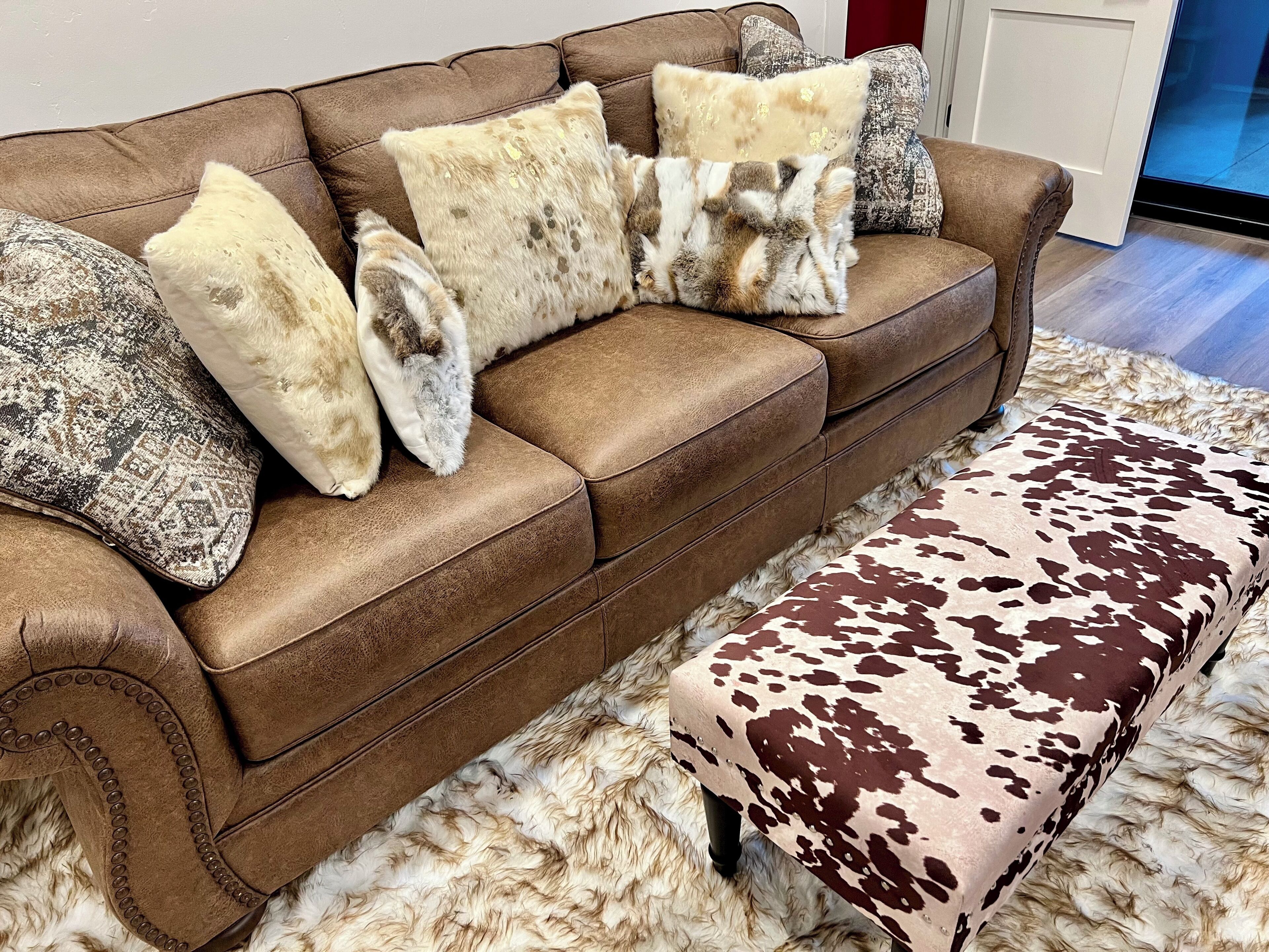 A brown leather sofa with patterned and fur-like cushions, a matching ottoman with a cowhide pattern, and a fluffy white rug.