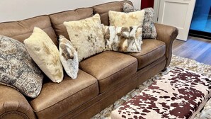 Ein braunes Ledersofa mit gemusterten und fellartigen Kissen, ein passender Ottoman mit Kuhfellmuster und ein flauschiger weißer Teppich.