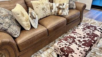 A brown leather sofa with patterned and fur-like cushions, a matching ottoman with a cowhide pattern, and a fluffy white rug.