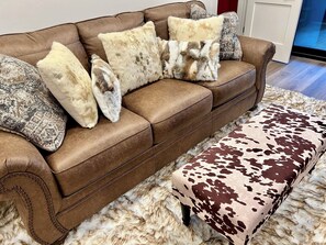 Un canapé en cuir marron agrémenté de coussins à motifs et à l’aspect fourrure, un pouf assorti avec un motif en peau de vache, et un tapis blanc et moelleux.
