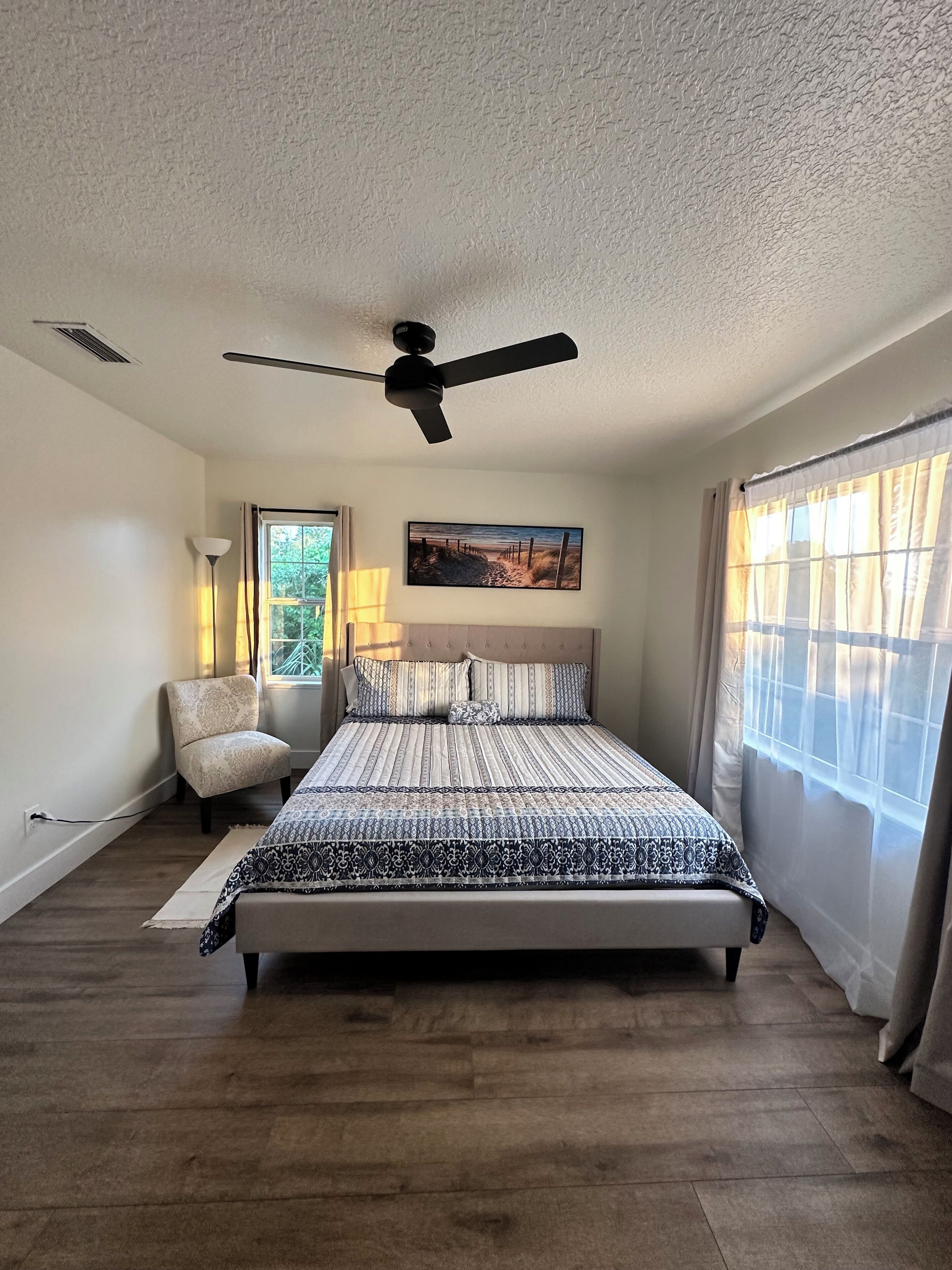 A bedroom with a large bed, a ceiling fan, a chair, and a window with curtains.
