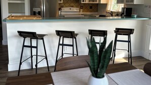 A modern kitchen with a central island, bar stools, a hanging light fixture, and a potted plant.