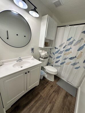 Un baño con lavamanos, espejo y retrete blancos. Hay una cortina de ducha con estampado de peces.