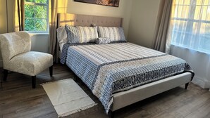 A bedroom with a bed, a chair, a window with curtains, a lamp, and a painting on the wall.