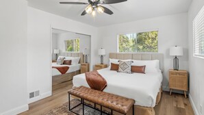 A bedroom with a large bed, a ceiling fan, a window with blinds, a bedside table with a lamp, and a bench. - Retreat home tucked away in the forest, perfect for big groups, near Bass Lake (Oakhurst)